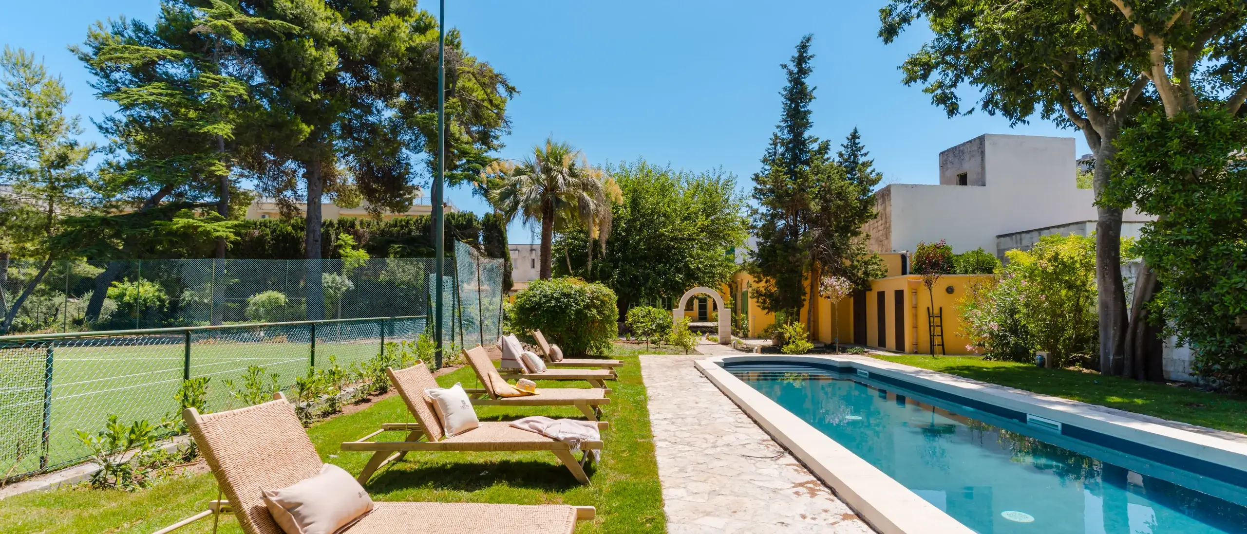 Una piscina circondata da lettini prendisole e alberi, con un campo da tennis visibile sullo sfondo