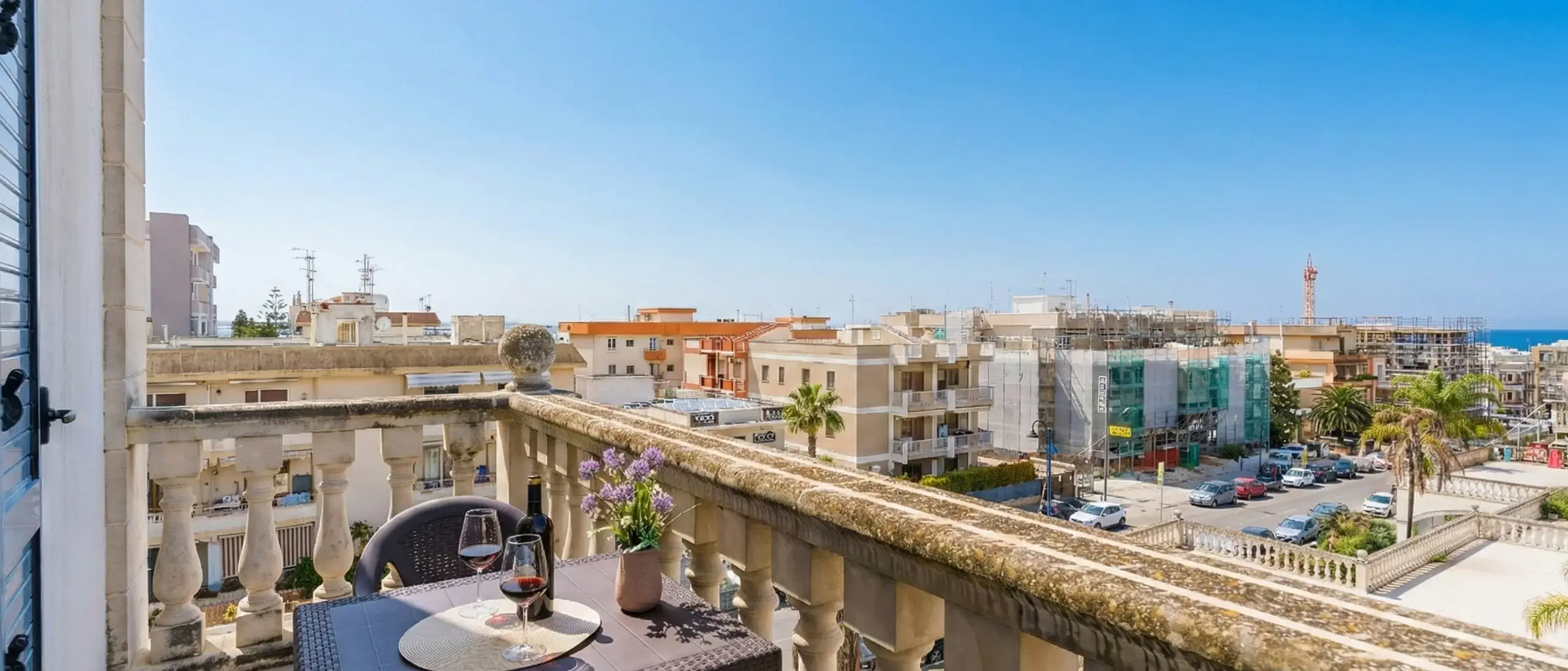 Vista da un balcone con un tavolo, due bicchieri di vino rosso e un vaso di fiori, che mostra edifici e il mare sullo sfondo