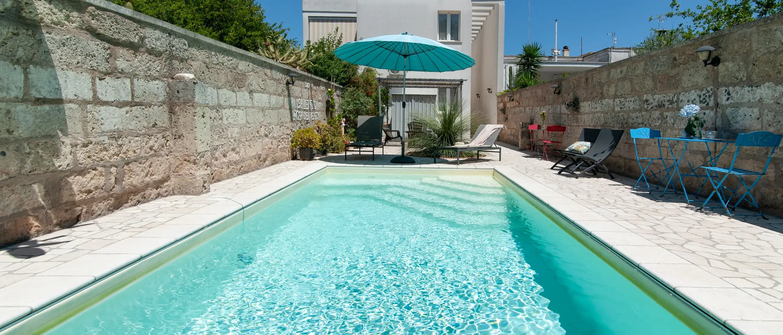 Una piscina circondata da pareti in pietra, con lettini, ombrellone e tavolo blu, in un ambiente soleggiato