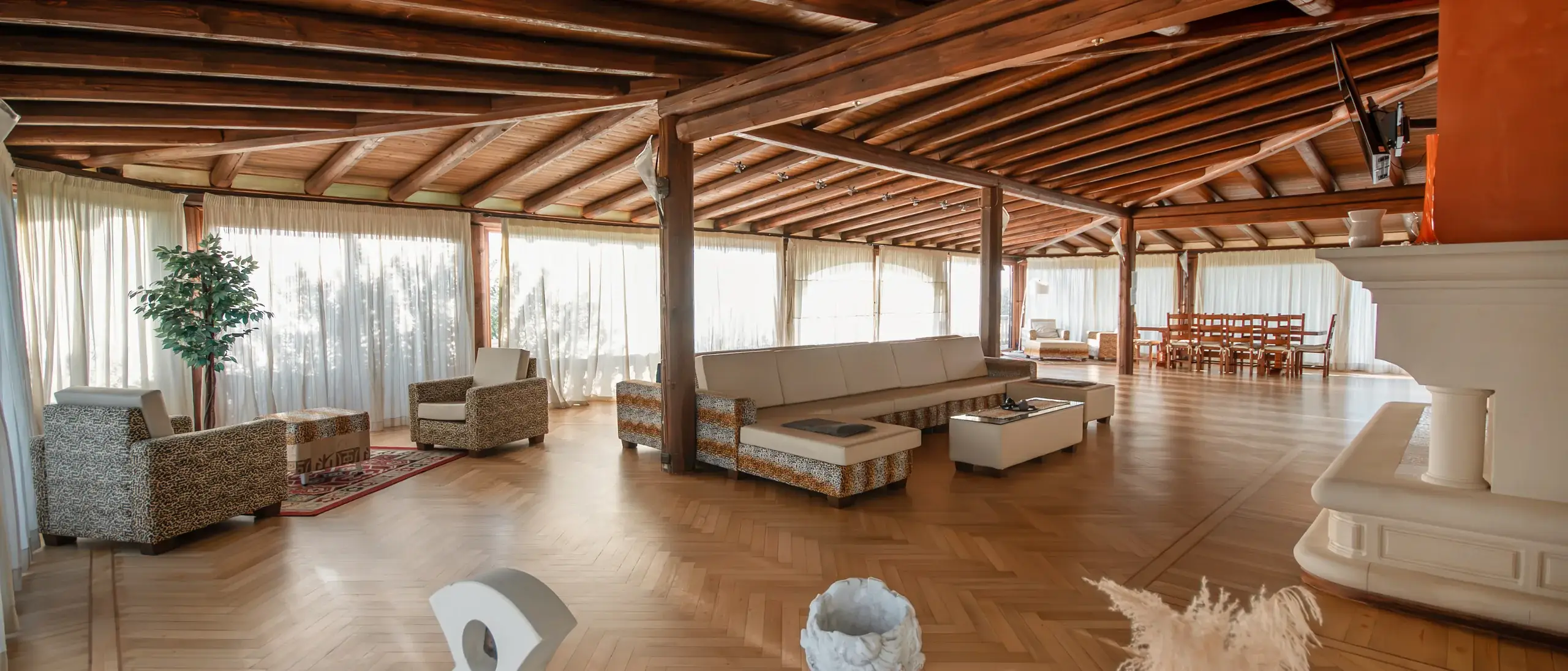 Spazio abitativo luminoso con soffitto in legno, divani eleganti e tavolo da pranzo, circondato da tende trasparenti