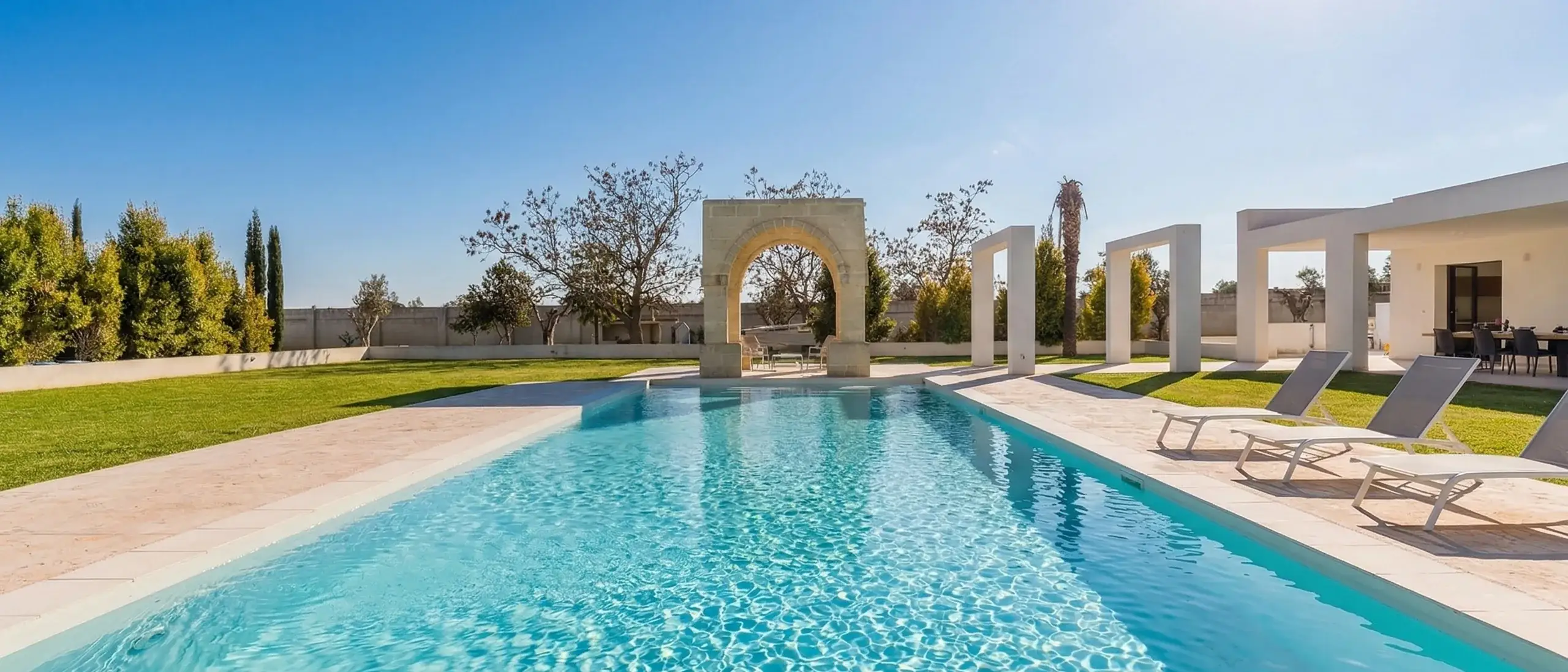 Una piscina cristallina circondata da un giardino ben curato, con un arco in pietra e sedie a sdraio