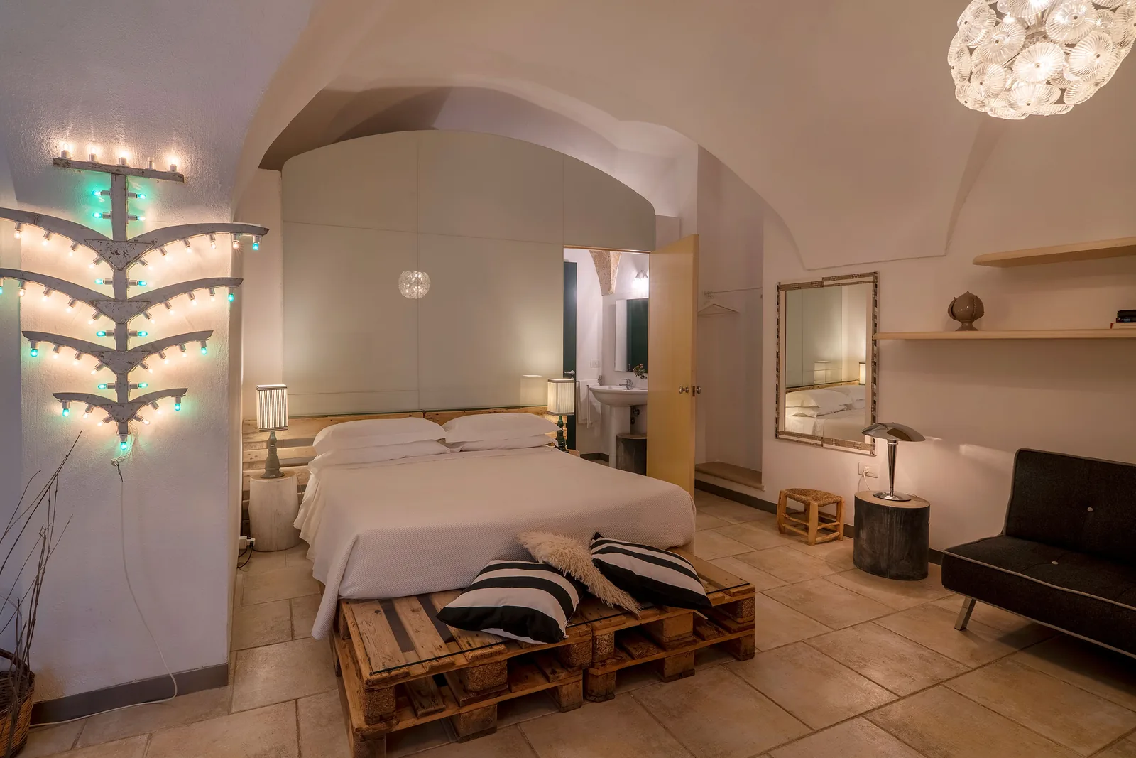 Camera da letto con letto su pallet, illuminazione decorativa, specchio e angolo soggiorno