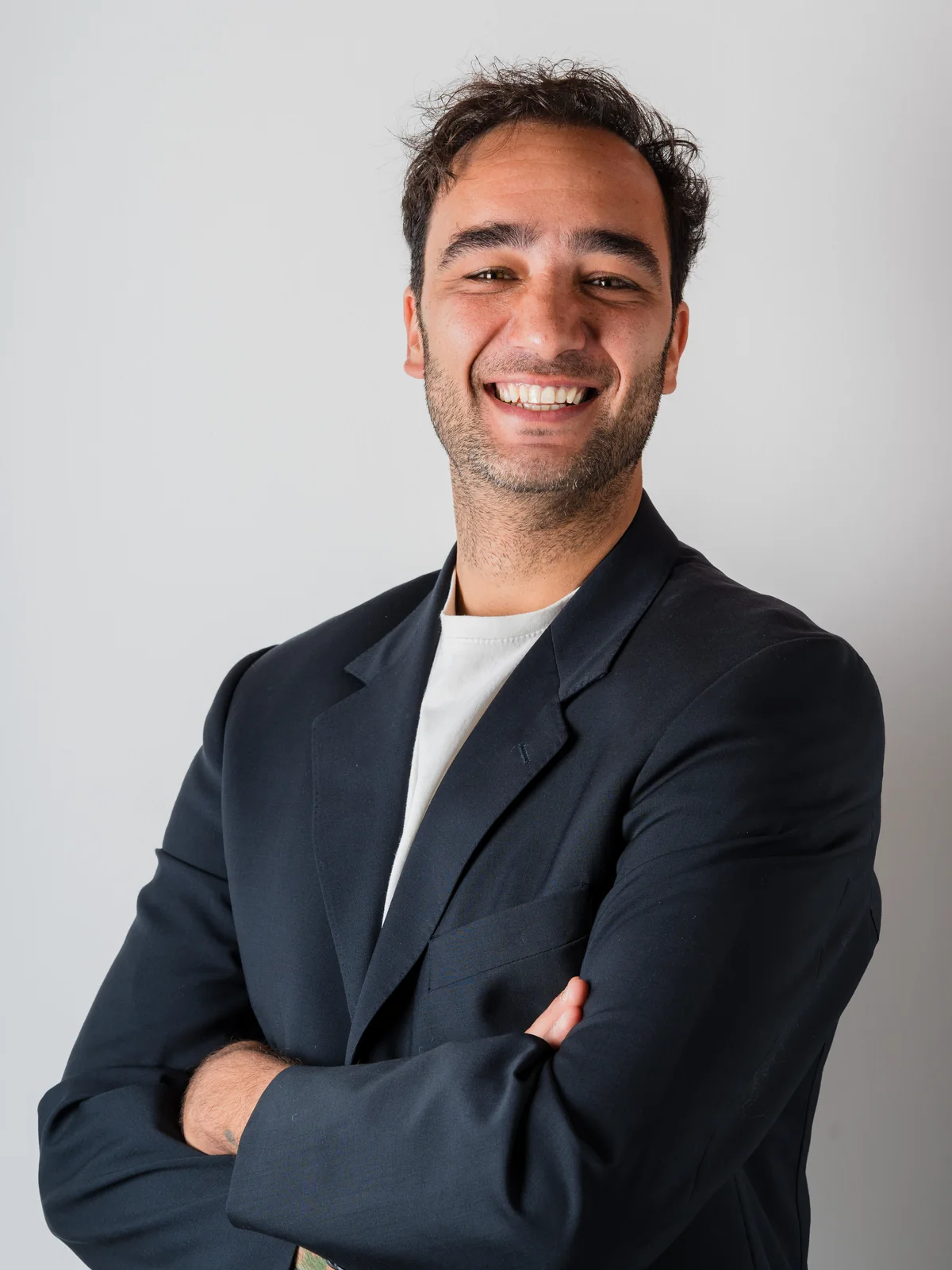 Un uomo sorridente con le braccia incrociate, indossa un blazer scuro e una maglietta bianca, su uno sfondo chiaro
