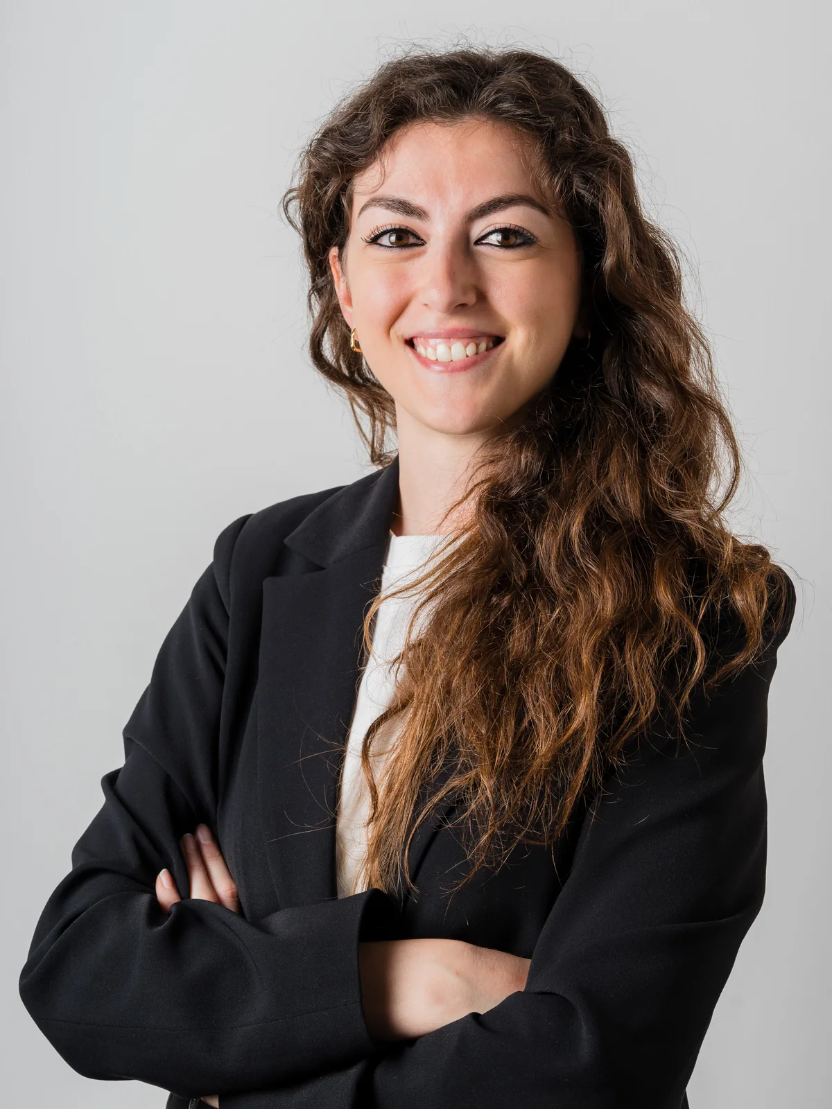 Una giovane donna sorridente con i capelli ricci e scuri, indossa un blazer nero e ha le braccia incrociate
