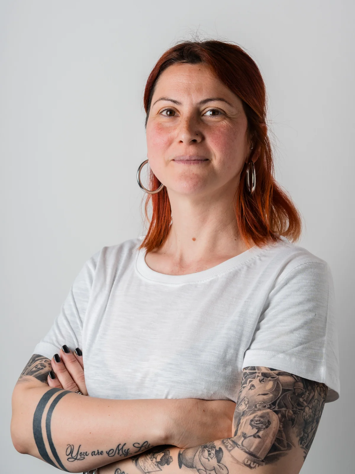 Una donna con capelli rossi e tatuaggi visibili sulle braccia, che indossa una maglietta bianca e ha un'espressione sicura
