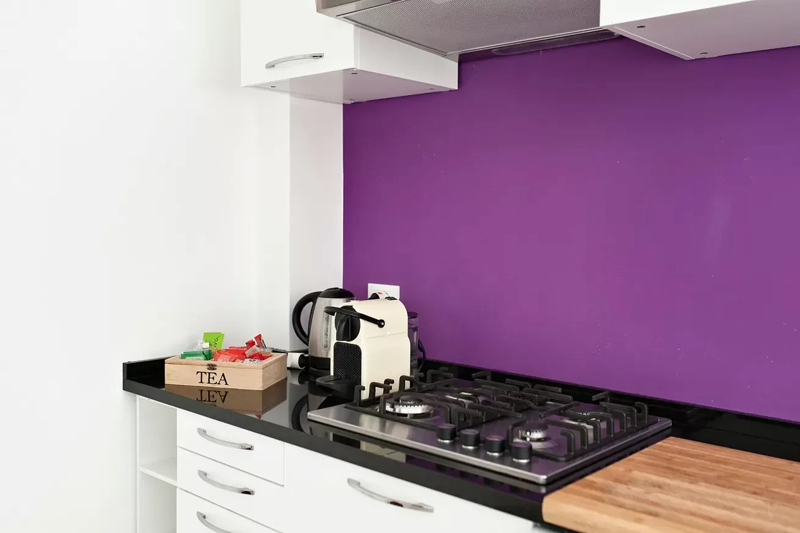 Un angolo di cucina con parete viola, piano cottura in acciaio, bollitore e un vassoio con bustine di tè