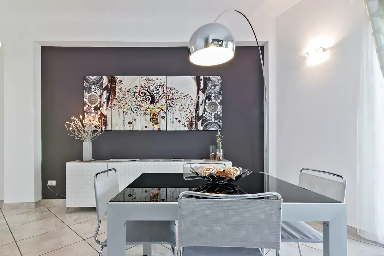 Un tavolo da pranzo moderno con sedie, illuminato da una lampada a sospensione, di fronte a un'opera d'arte colorata su un muro grigio