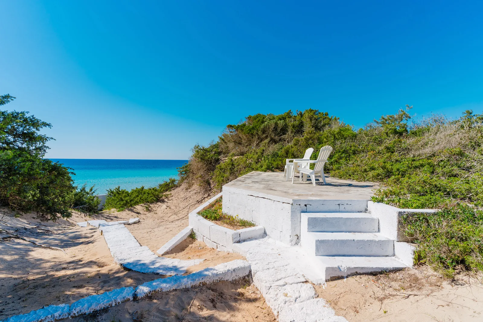 Un'area di relax con due sedie bianche su una piattaforma, circondata da vegetazione e con vista sul mare azzurro