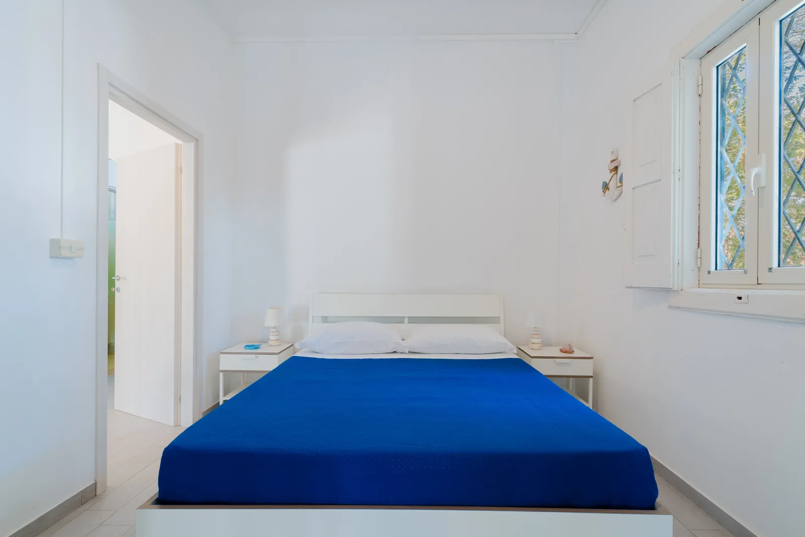 Camera da letto luminosa con letto matrimoniale e biancheria blu, armadietti bianchi e una finestra