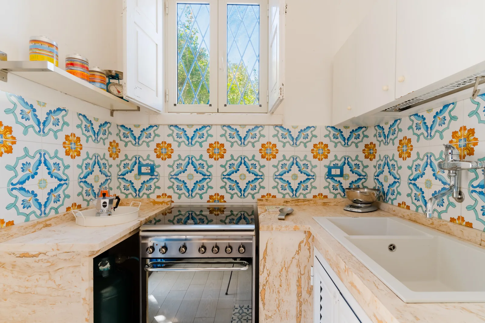 Cucina con piastrelle decorative blu e arancioni, piano cottura in acciaio inox e lavello bianco. Finestra aperta con vista verde