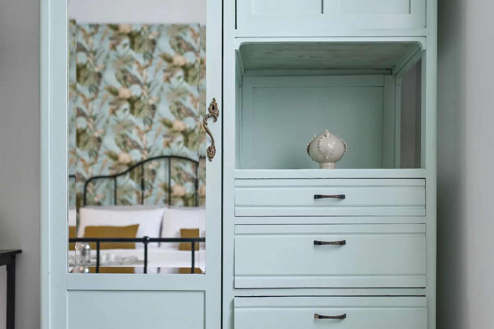 Un mobile azzurro con un vaso decorativo all'interno, riflettendo un letto con cuscini gialli e una carta da parati floreale sullo sfondo