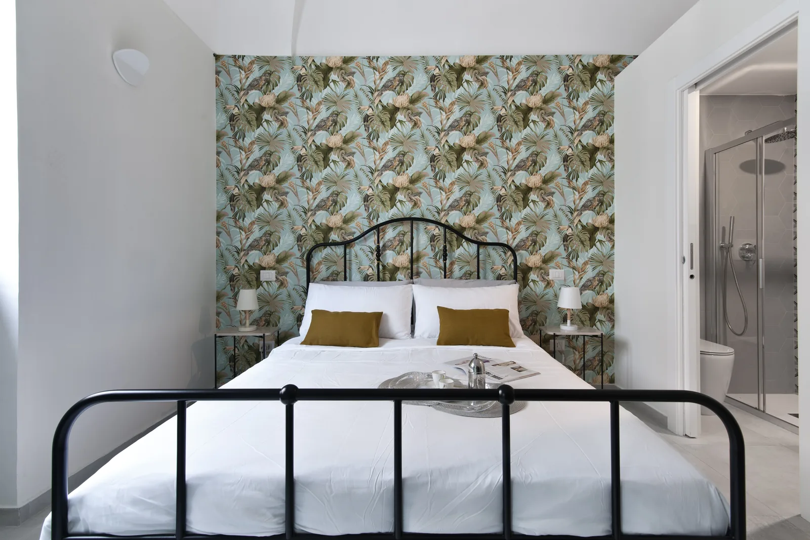 Camera da letto con parete decorata da carta da parati floreale, letto matrimoniale con cuscini gialli e bagno visibile