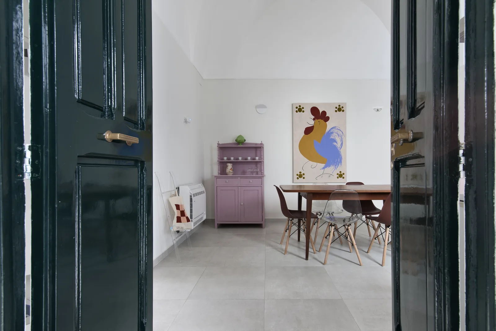 Vista di una sala da pranzo con un tavolo in legno, sedie moderne e un armadietto viola, decorata con un dipinto di un gallo