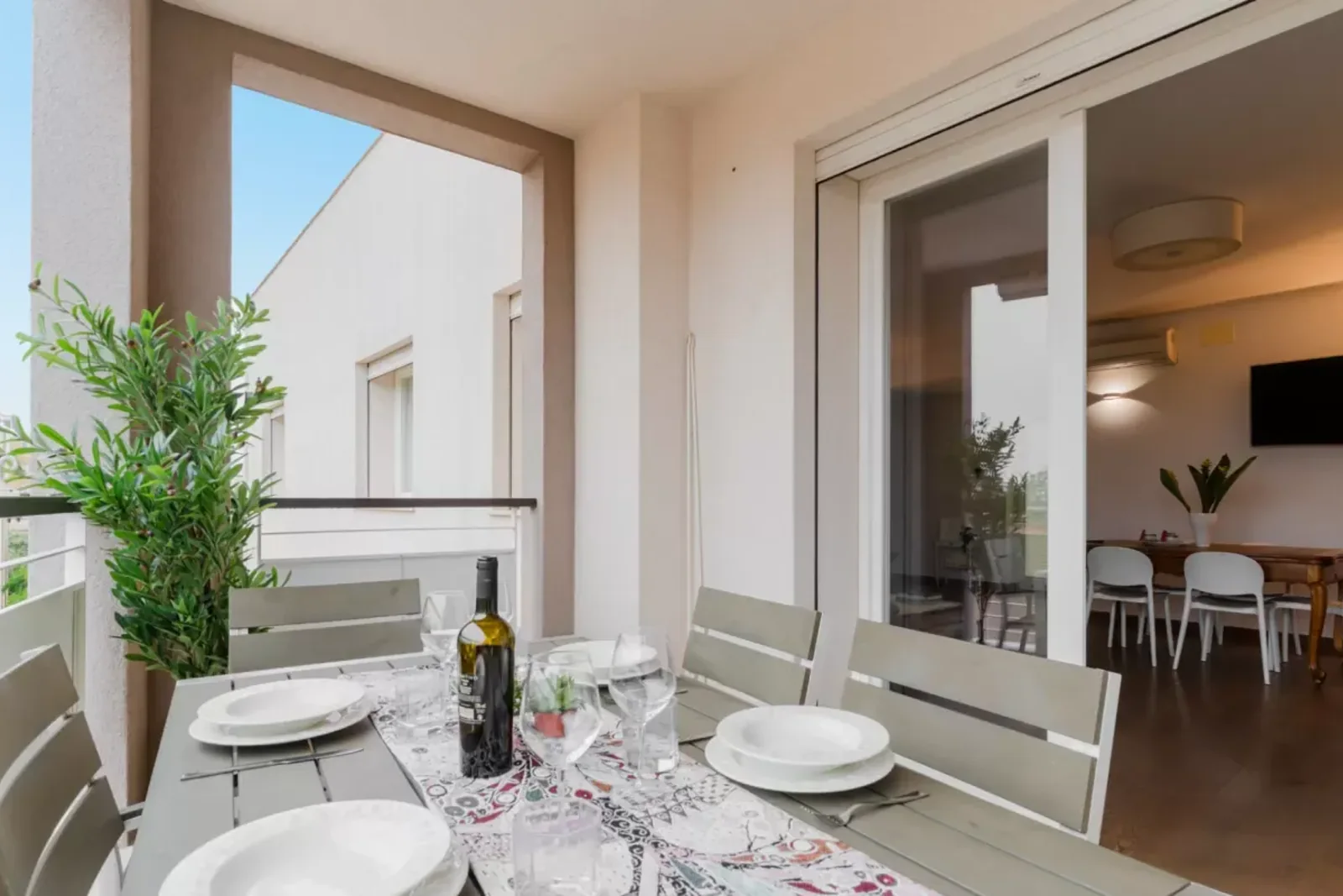 Un tavolo apparecchiato su un balcone, con piatti, bicchieri e una bottiglia di vino, affacciato su un interno luminoso