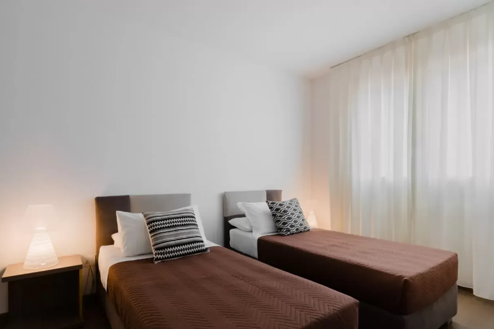 Due letti singoli con copriletto marrone e cuscini decorativi, lampada accesa su un comodino, tenda bianca