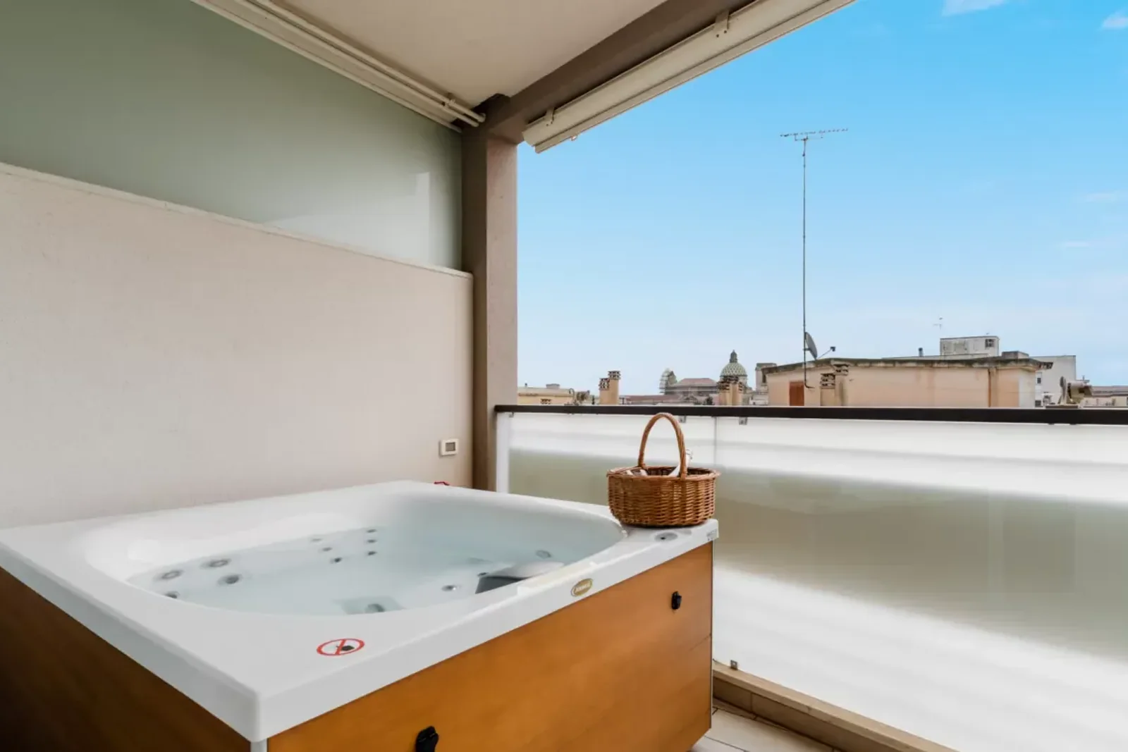 Vasca idromassaggio su un balcone con vista sui tetti della città, accompagnata da un cesto di vimini