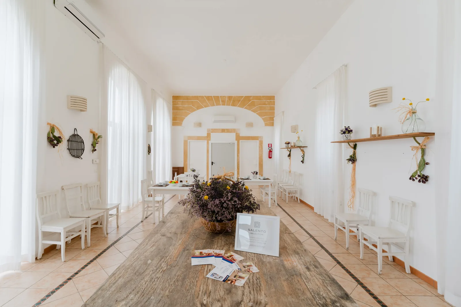 Una sala luminosa con tavolo in legno, sedie bianche e decorazioni floreali. Un volantino su un tavolo presenta informazioni su Salento