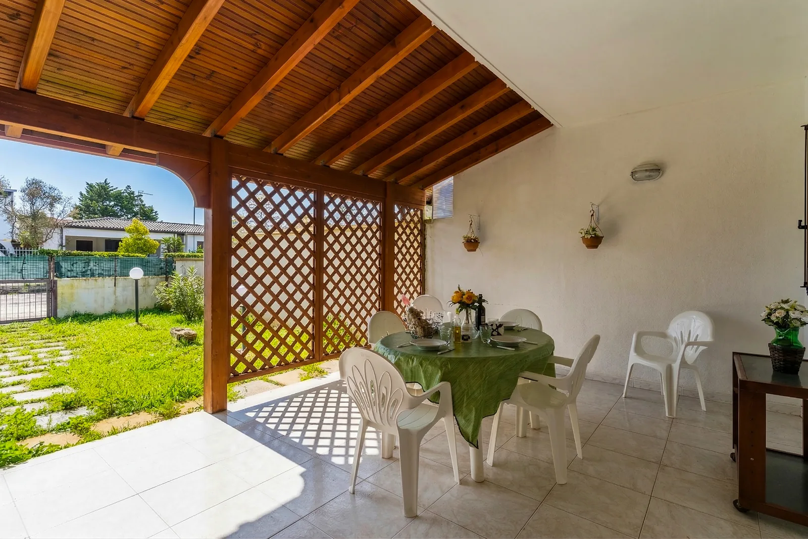 Un'area esterna coperta con un tavolo apparecchiato e sedie bianche, circondata da un giardino verde e una recinzione