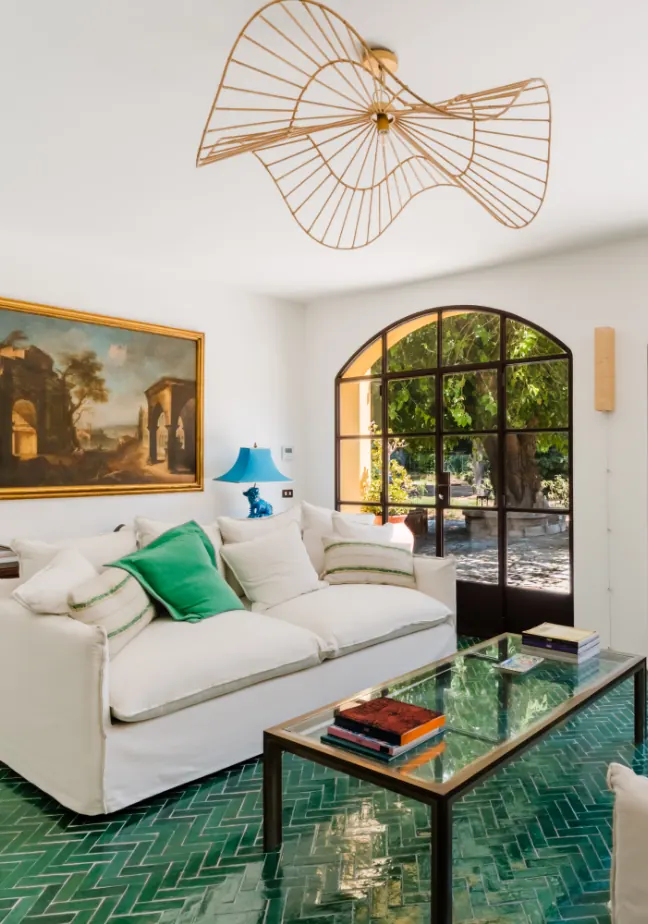 Un soggiorno luminoso con un divano bianco, cuscini verdi, un tavolino di vetro e una lampada blu, con una grande finestra che mostra il giardino