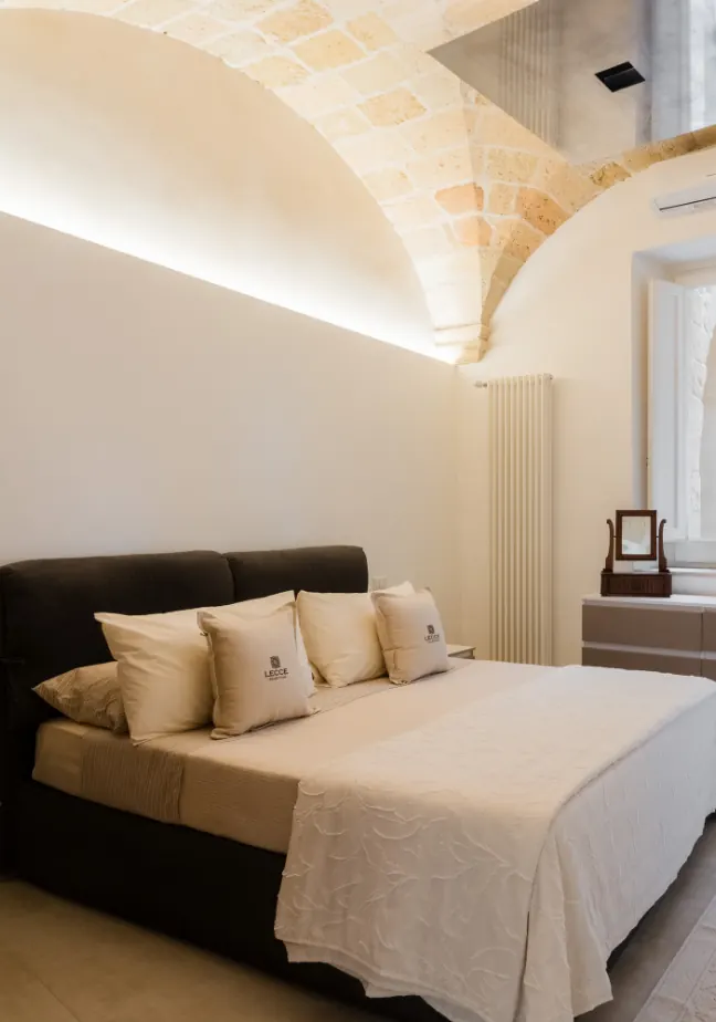 Camera da letto moderna con un letto matrimoniale, cuscini con il logo "Lecce" e un arredamento minimalista